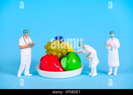 Three miniature doctor figurines in white lab coats and face masks examine a small white plate of plastic fruit, including a strawberry and grapes, on Stock Photo