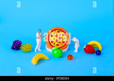 Miniature surgeons in scrubs examine a toy pizza on a blue surface surrounded by plastic fruit models including grapes, banana, apple, and strawberry. Stock Photo