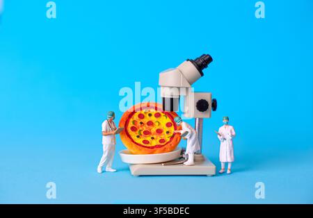 Miniature scientists conducting food quality analysis on a pizza using a microscope against a vivid blue background, illustrating concepts of food saf Stock Photo