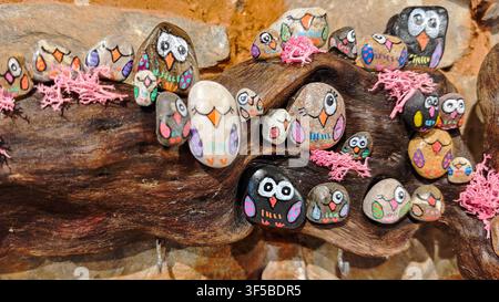 Close-up of decorative pebble art featuring colorful hand-painted stone owls perched on a rustic piece of wood with pink moss. Souvenir craft. Stock Photo