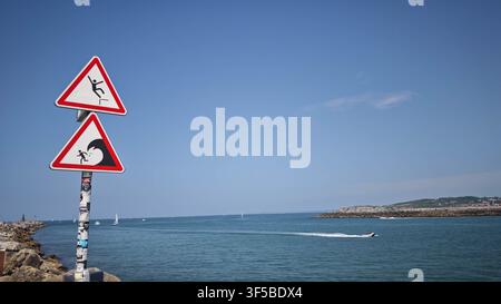 Close-up of funny red danger signs on a sea pier showing a person falling and a wave hazard. Clear turquoise ocean and blue sky background. Stock Photo