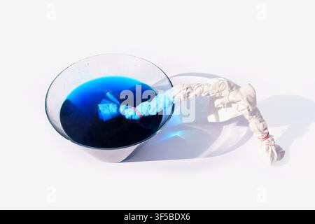 Tie dye process showing a rolled white cotton fabric tied with rubber bands partially dipped in a glass bowl filled with vibrant blue liquid dye on wh Stock Photo