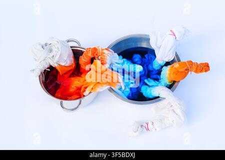 Tie dyeing process showing white fabric tied with rubber bands being dipped into two metal pots filled with bright orange and deep blue liquid dyes on Stock Photo