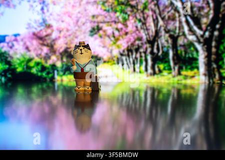 Chiang Mai, Thailand - 30 July 2026 : Traveler cat figurine wearing suit holding suitcase reflected on silver surface, cherry blossom trees background Stock Photo
