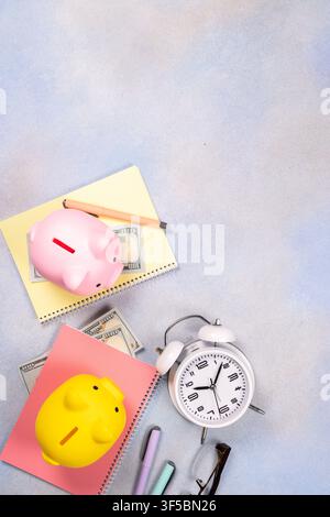 Two colorful piggy banks, cash, notebooks, pens, glasses, and an alarm clock on a textured light background, representing financial planning and savin Stock Photo