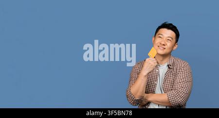 Thoughtful Asian Man Holding Credit Card And Smiling, Concept Of Online Shopping Stock Photo