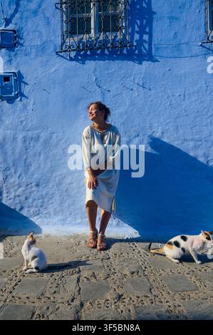 Chefchaouen, Morocco, 30th of July 2026: A woman in a white dress relaxes against a vivid blue wall while two cats lounge on a sunlit cobblestone stre Stock Photo