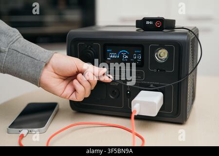 Man plugging cable into portable power station charging devices at home during blackout  Stock Photo