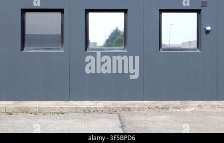 Prefab gray concrete wall with a row of large windows reflecting the industrial area around. Sidewalk and road in front. Background for copy space. Stock Photo