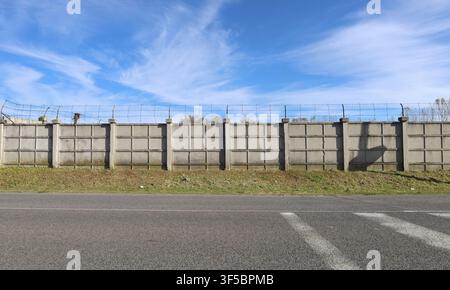 Long prefab concrete wall with barbed wire on top. Sky above, strip of grass and road in front. Background for copy space. Stock Photo