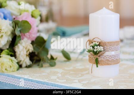 Rustic white candle decorated with burlap, lace, and small flowers on a floral wedding reception table.Elegant country-style wedding reception layout, botanical table setting Stock Photo