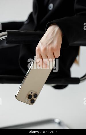 Gdansk, Poland - June 29, 2023: Businesswoman sitting on a chair holding a metallic smartphone. Professional executive using premium mobile phone, corporate workspace technology Stock Photo