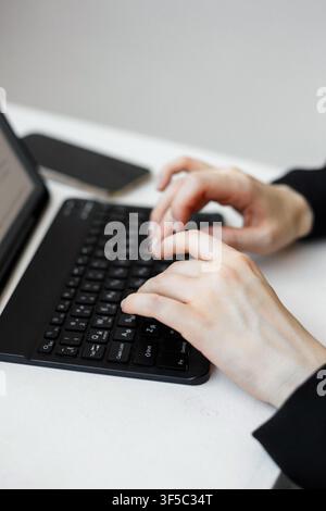 Close-up of female hands typing on a black portable keyboard with a tablet computer on white desk. Wireless smart device workspace, remote freelance blogging, digital nomad lifestyle Stock Photo