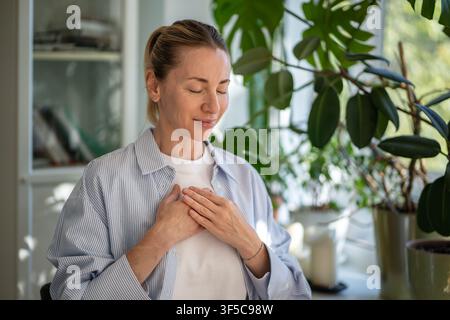 Relaxed smiling woman meditates with hands on chest, practice breathing exercises at home. Self care Stock Photo