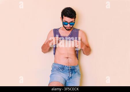 Young man wearing sunglasses lifting his shirt, feeling the heat.  Man in summer attire, fisherman wearing a straw hat. Young man in a blue Stock Photo