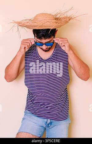 Young man taking off sunglasses. Young male tourist wearing a striped sailor-style tank top and short denim shorts; feeling the summer heat Stock Photo
