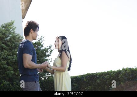 Couple standing holding hands near trimmed hedge under clear sky, wearing navy, yellow clothes Stock Photo