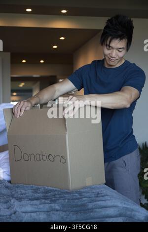 Asian adult male wearing navy T-shirt packing donations box on bed with gray blanket Stock Photo