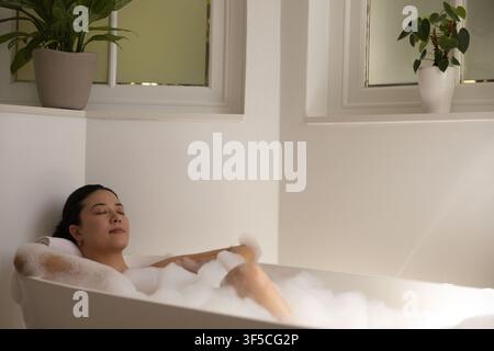 African American woman reclining and bathing in standalone tub with bubble foam and frosted windows Stock Photo