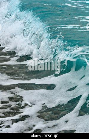 A close-up action shot of a turquoise ocean wave crashing and breaking over smooth pebbles on a coastal beach, creating white sea foam and splashing water droplets. Stock Photo