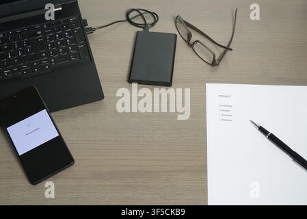 High-angle shot of a workstation including a laptop, "Project" checklist, external hard drive, and a phone displaying a successful backup. Stock Photo