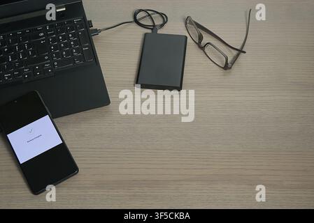 Minimalist desk view showing a laptop connected to an external hard drive and a smartphone with a "Backup Complete" message. Stock Photo