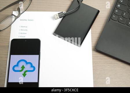 Smartphone displaying a cloud icon resting on a "Digital Tasks" checklist with an external drive and USB cable. Stock Photo