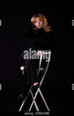 Young woman sitting on a white chair, looking pensive with a dark background Stock Photo