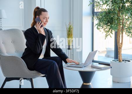 Businesswoman talking on phone and using laptop in modern bright office Stock Photo