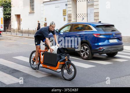 A man riding an orange cargo bike along a city street with a blue SUV in the background Stock Photo