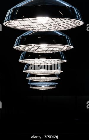 Row of illuminated industrial style pendant lamp fixtures with metal grid guards hanging in perspective against a dark background Stock Photo