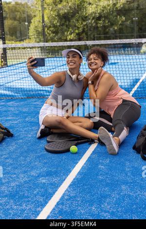 Two African American women sitting on blue court in sportswear taking selfie with phone, paddles. Pickleball, outdoor, duo, friendship, fitness, headphones, backpacks Stock Photo