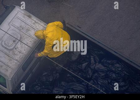Worker in yellow raincoat loading black garbage bags into truck Stock Photo