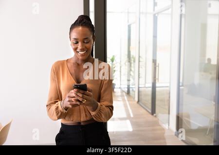 Smiling businesswoman texting on smartphone standing in office hallway Stock Photo