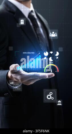 A businessman in a dark suit presents a futuristic virtual interface displaying data analysis, growing bar charts, risk indicators, and financial icon Stock Photo