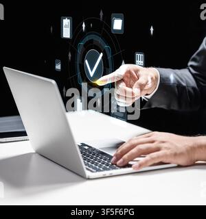 Businessman using a laptop while interacting with a digital holographic interface featuring a checkmark, symbolizing data verification, security, qual Stock Photo