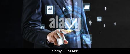A professional businessman pointing at a glowing blue checkmark icon on a virtual dashboard representing quality assurance, certification, and success Stock Photo