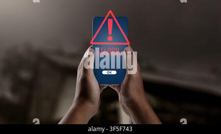 A hand holds a smartphone showing a flood warning alert, with an overturned car partially submerged in water under a dark, stormy sky in the backgroun Stock Photo