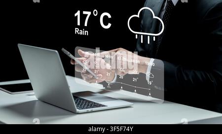 A businessman in a suit uses a laptop while a futuristic transparent digital interface displays a detailed weather forecast with temperature and rain Stock Photo