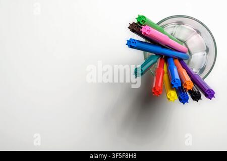 Top-down view of vibrant multi-colored felt-tip pens in a clear glass on a white surface, featuring ample copy space on the left for text Stock Photo