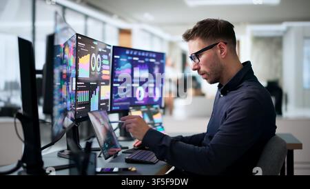 Young Man Uses A Laptop And Tablet To Track Business Performance Data. Stock Photo