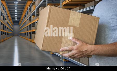 Close-up of a warehouse worker carrying a sealed cardboard shipping box in a modern distribution center with storage racks, representing logistics, fu Stock Photo