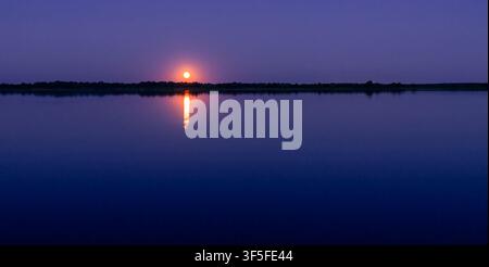 Full Moon Rising Above Calm Lake with Bright Reflection During Peaceful Blue Twilight Stock Photo