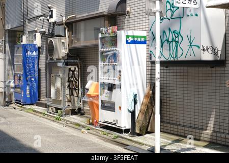 A white Ito En vending machine and a blue Asahi vending machine with graffiti standing along a narrow alleyway in Tokyo, Japan. Stock Photo