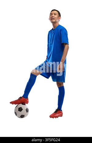 A young Indonesian southeast asian male athlete in a full blue soccer kit stands confidently with one foot resting on a soccer ball, isolated against Stock Photo
