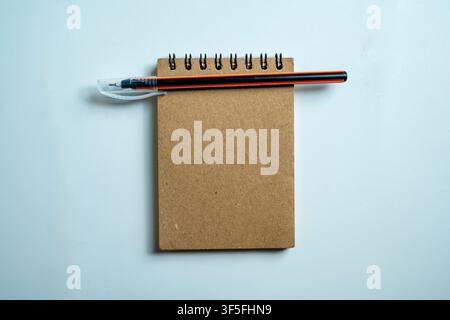 A top-down view of a blank brown spiral-bound notepad with an orange and black pen resting on it, set against a neutral gray textured background Stock Photo