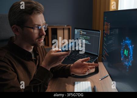 Close up of a man wearing glasses talking to an AI chatbot on a smartphone while working at a computer with artificial intelligence agent. Man using artificial intelligence assistant for work.  Stock Photo