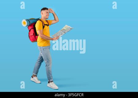 Happy millennial tourist with tourist gears and map looking ahead while walking on blue studio background, copy space Stock Photo