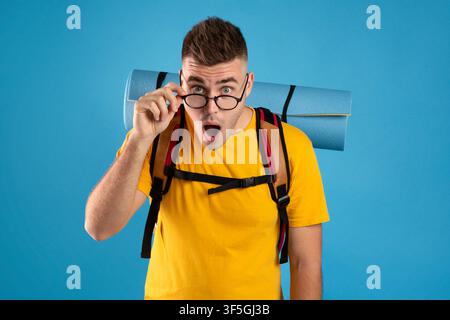 Shocked young guy with tourist equipment staring at camera with open mouth, blue studio background Stock Photo