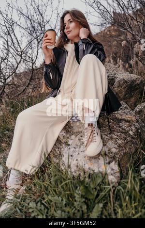 Woman coffee outdoors woman sits on a rock holding a paper coffee cup while relaxing in a mountainous landscape during daytime Stock Photo